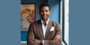 Confident professional standing by a window with arms crossed, wearing a brown blazer and white shirt in a modern office.