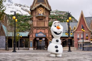 Smiling snowman character Olaf stands in front of a Frozen-themed clock tower and village buildings at a winter-inspired theme park area.