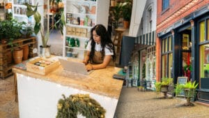Small business owner working on a laptop inside a plant and garden shop, with a split view of a colorful brick storefront on a downtown street.