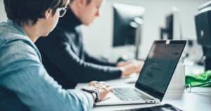Two people working at a desk on laptop computers in a modern office.