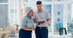 Two coworkers standing in a bright modern office reviewing information together on a digital tablet