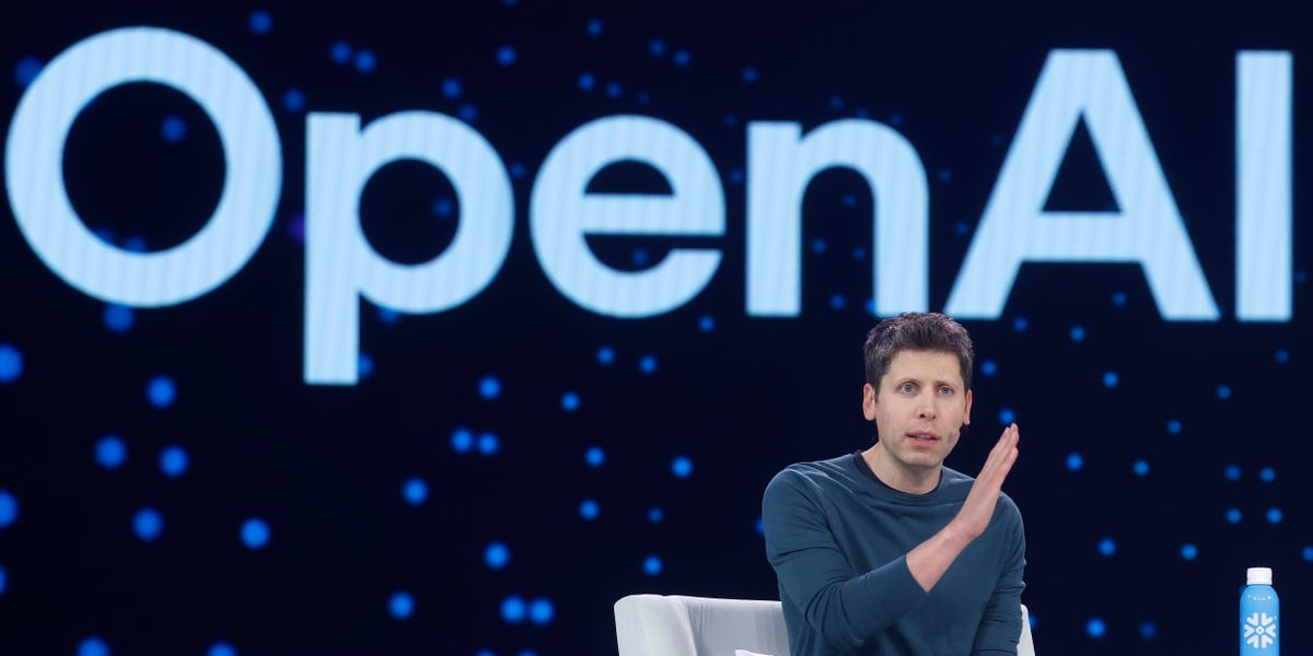 A speaker sitting on stage in front of a large OpenAI logo, gesturing with one hand during a presentation.