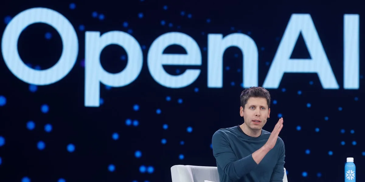 A speaker sitting on stage in front of a large OpenAI logo, gesturing with one hand during a presentation.
