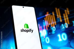 Shopify logo on a smartphone screen with digital financial charts in the background