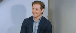 Smiling person wearing a blazer and plaid shirt, seated indoors during a conversation